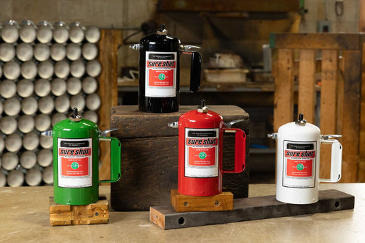 A display of four Sure Shot A1000 spray canisters arranged on tiered wooden blocks, with green, red, and white canisters in the foreground and a black canister elevated on a crate in the background.