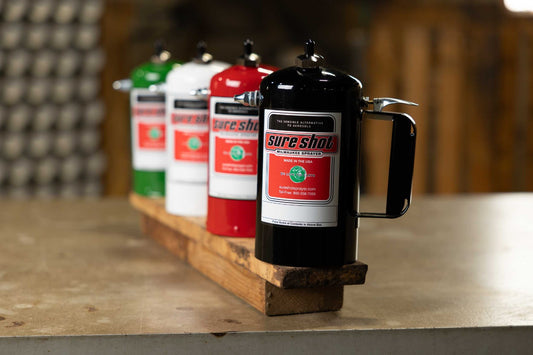 A black Sure Shot A3000 stainless steel spray canister in sharp focus in the foreground, with red, white, and green canisters lined up behind it on a wooden board in a workshop.